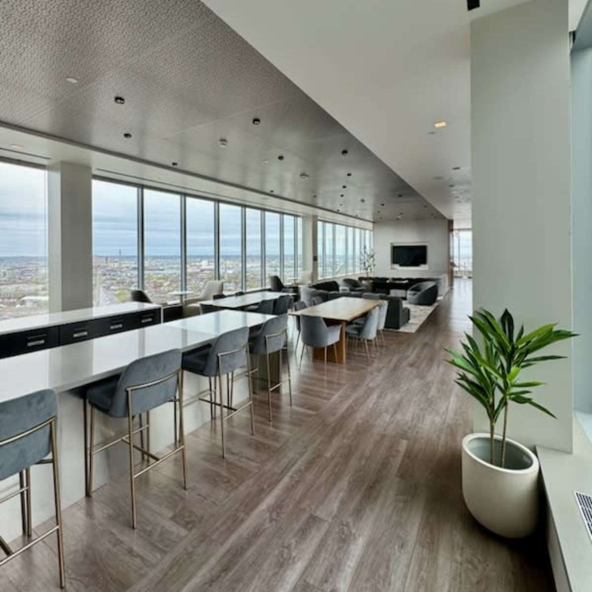 A modern office space with large windows, featuring a dining area with high stools, tables, and a lounge area with a television.