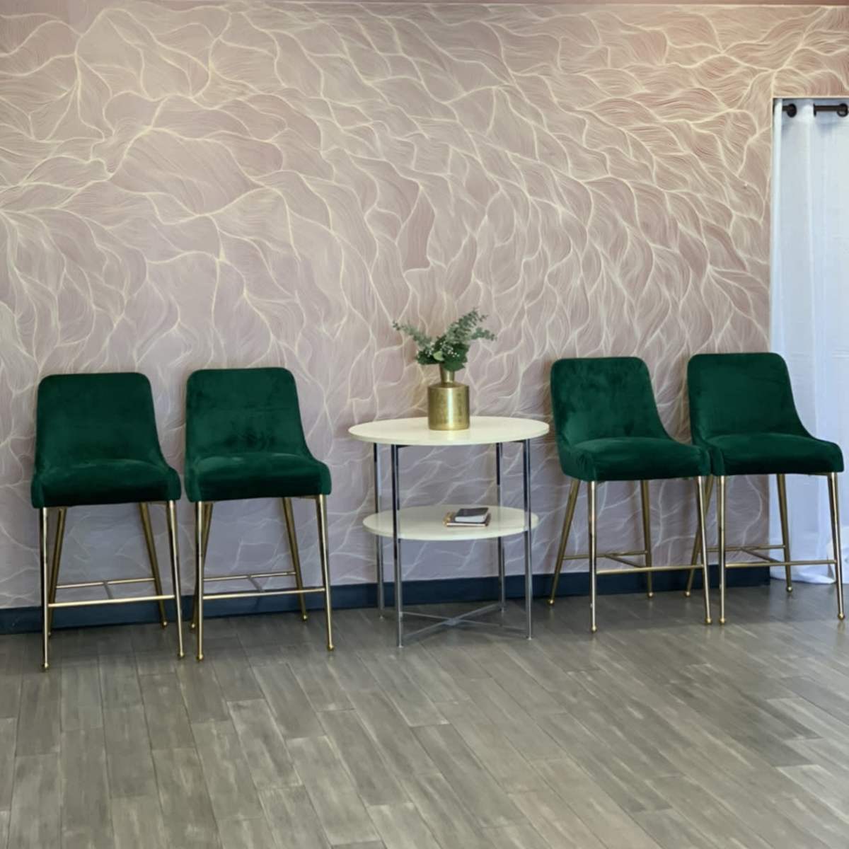 Four green velvet bar stools with gold legs arranged around a small round table on a wooden floor against a patterned wall.