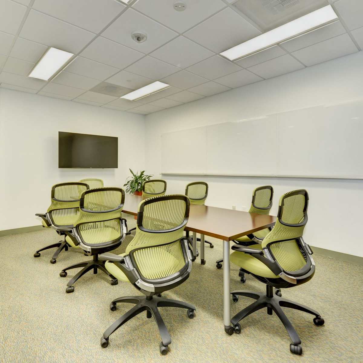 A conference room with a rectangular table, eight rolling chairs, a wall-mounted television, and a dry-erase board.