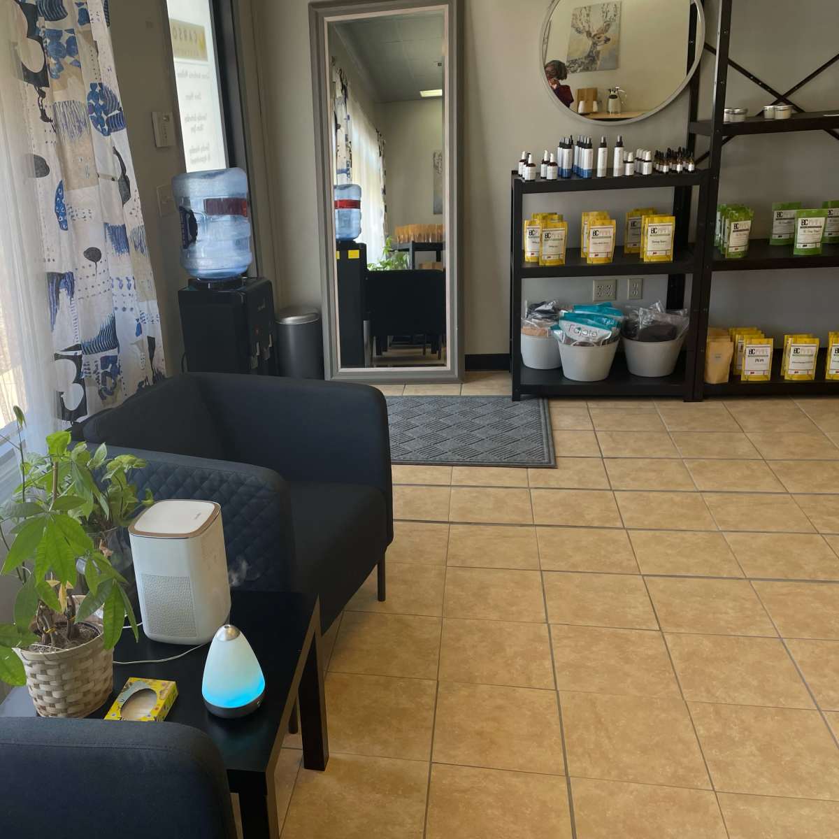 A waiting area with two black chairs, a small potted plant, a water cooler, a mirror, and shelves filled with containers.