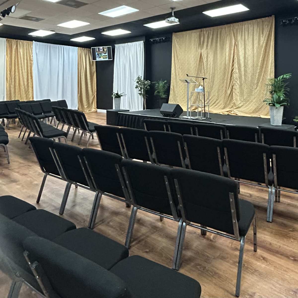 An auditorium-style room with black chairs arranged in rows facing a small stage decorated with white and gold drapes.