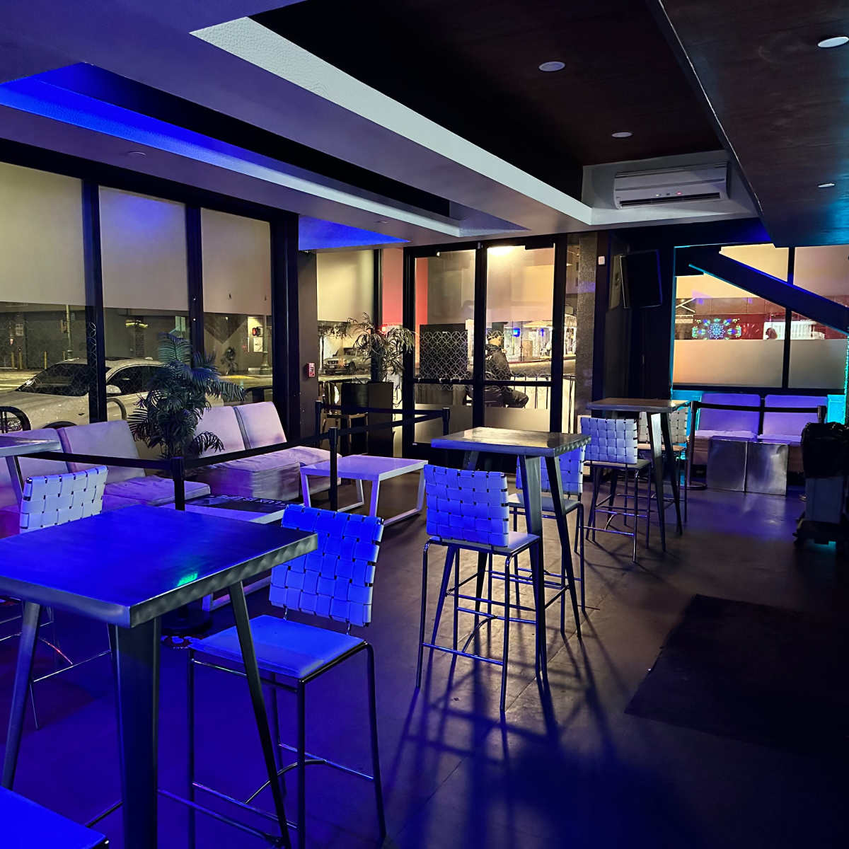 A dimly lit lounge area with several tall tables and chairs arranged around, and large windows revealing a street view outside.