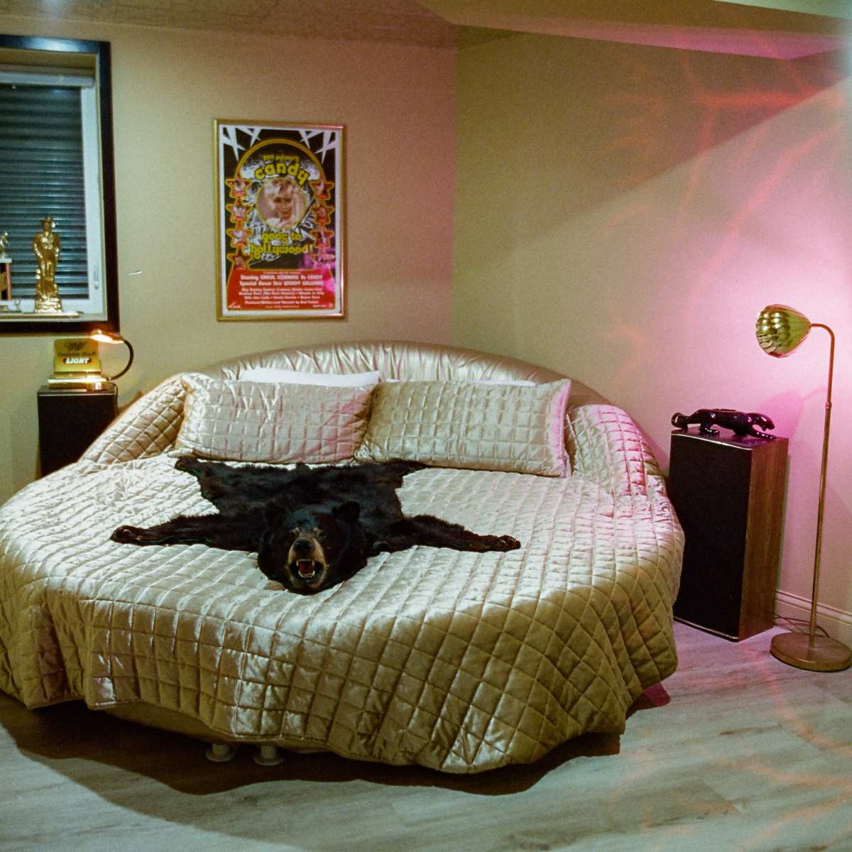 A round bed covered with a golden quilt is positioned in a basement room, featuring a mounted bear rug laid over it and soft lighting from a lamp and wall fixtures.