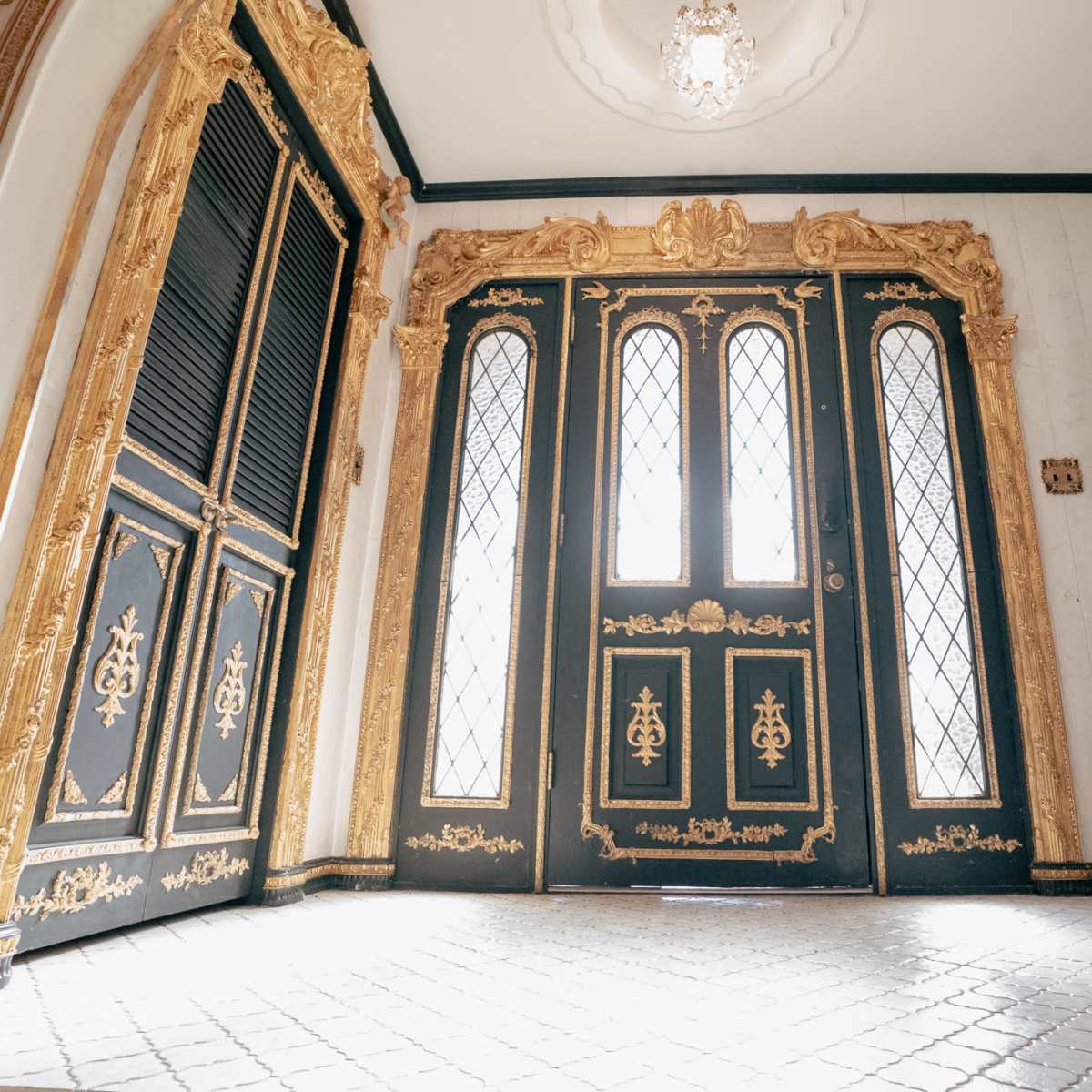 An ornate entrance with two large doors featuring intricate gold designs and diamond-patterned glass panels, illuminated by a chandelier above.