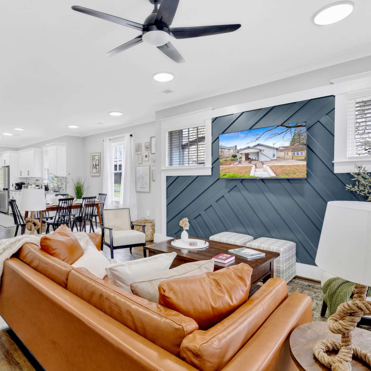 The living room features a brown leather sofa facing a mounted TV on a blue accent wall, with a dining area and kitchen visible in the background.