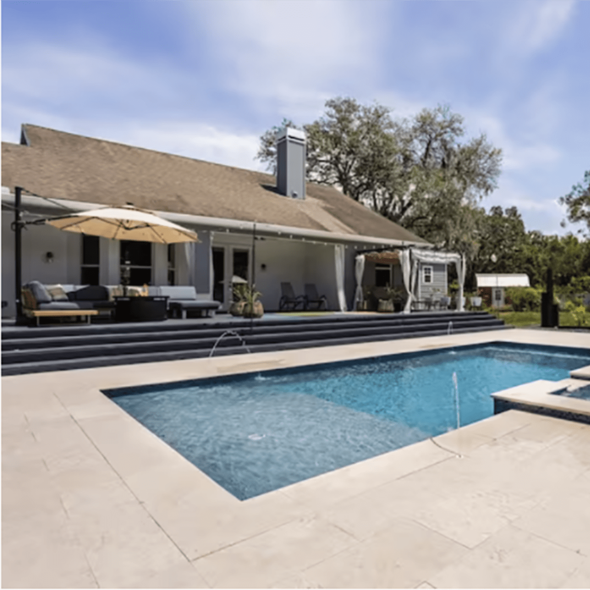 A backyard with a swimming pool, a hot tub, lounge chairs, and umbrellas, alongside a house and landscaped greenery.