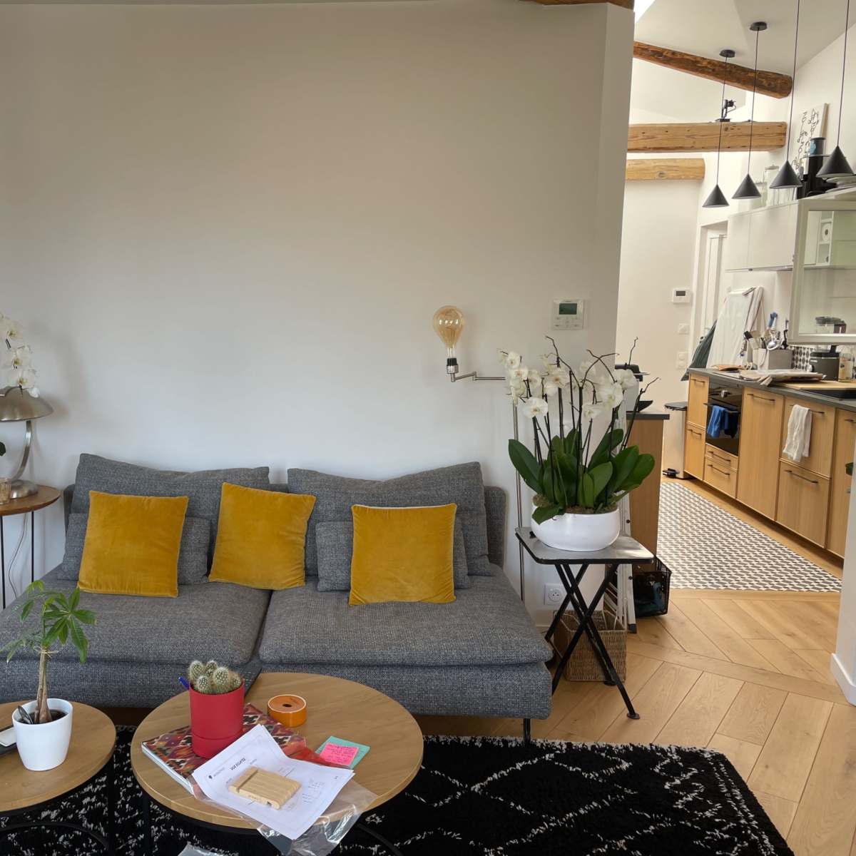 A cozy living area with a gray sofa adorned with yellow cushions, a wooden coffee table with various items, and a kitchen visible in the background.