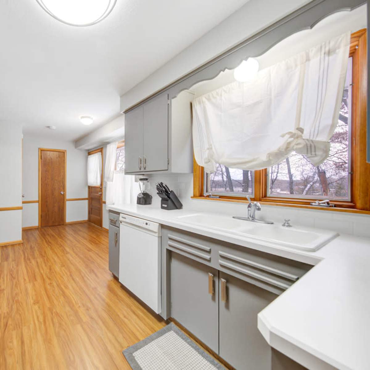 The kitchen features grey cabinetry, a white sink, and a window that provides natural light, with a door leading to another room in the background.