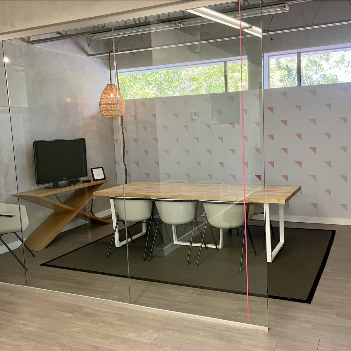 A modern conference room features a wooden table surrounded by white chairs, a television on the wall, and large glass windows with natural light.