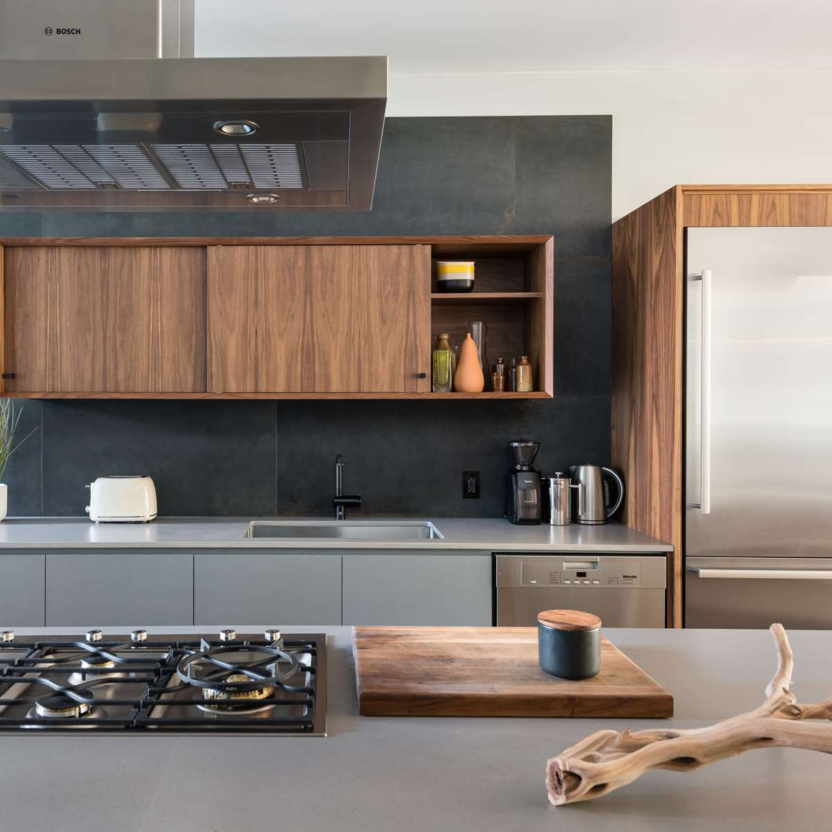 A modern kitchen featuring a gas cooktop, stainless steel appliances, wooden cabinetry, and a stone countertop.