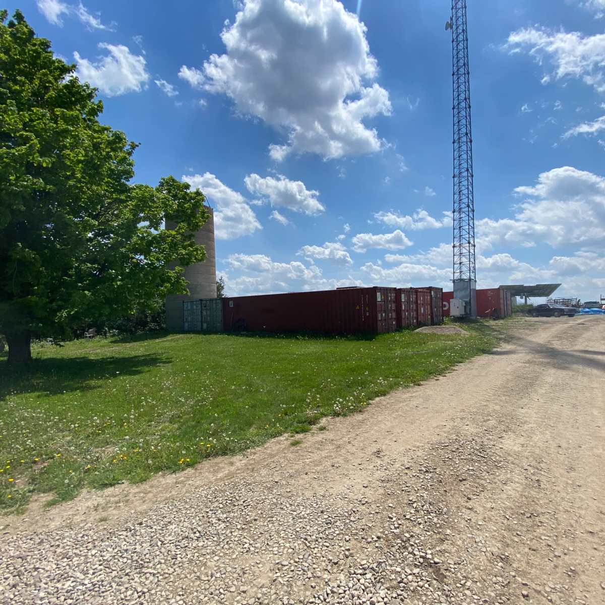 A dirt road leading past shipping containers and a tall tower under a partly cloudy sky.