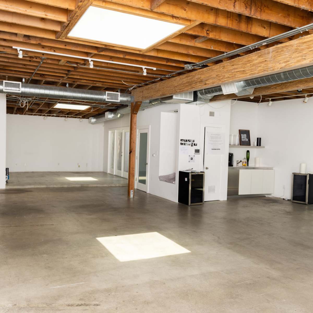 A spacious, minimalist interior with exposed wooden beams and concrete flooring, featuring a small furniture arrangement in one corner.