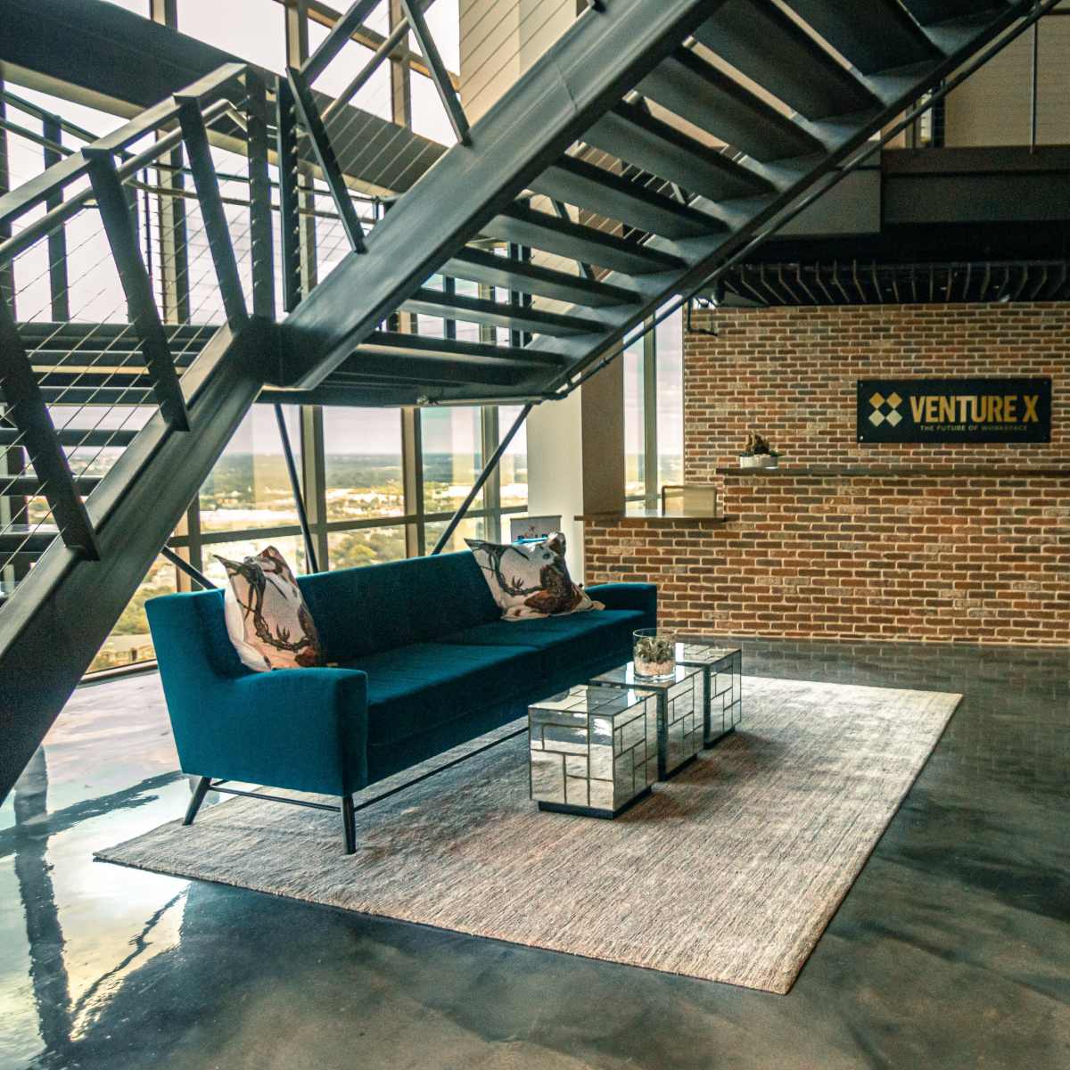 A modern office lobby featuring a blue sofa, glass coffee tables, and a brick accent wall with the "VENTURE X" logo.
