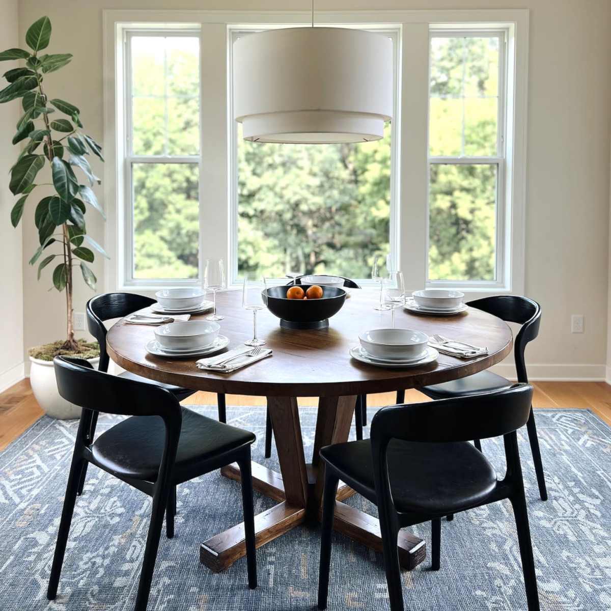 A round wooden dining table is set with four black chairs, a bowl of oranges in the center, and large windows providing a view of greenery outside.