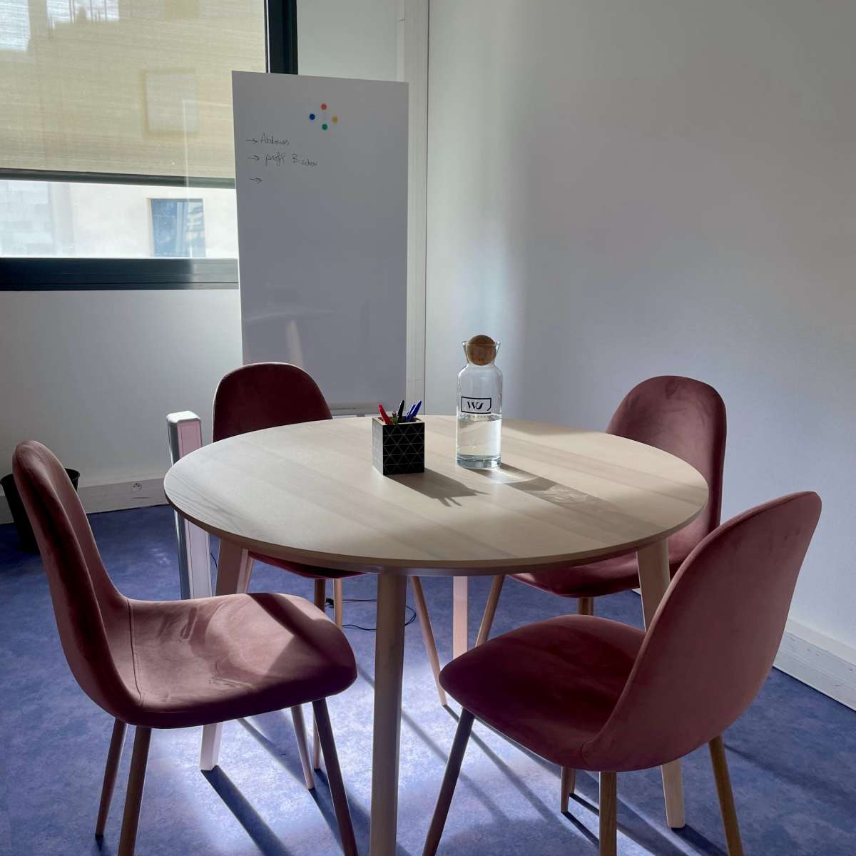 A round table with four pink chairs is set up in a bright office space next to a whiteboard.