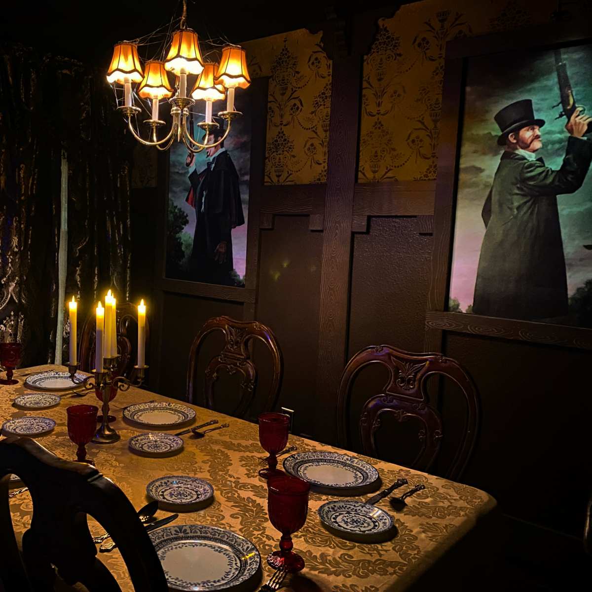 A dimly lit dining room features an elegant table set for dinner, adorned with patterned plates and red goblets, beneath a chandelier and framed portraits on the walls.
