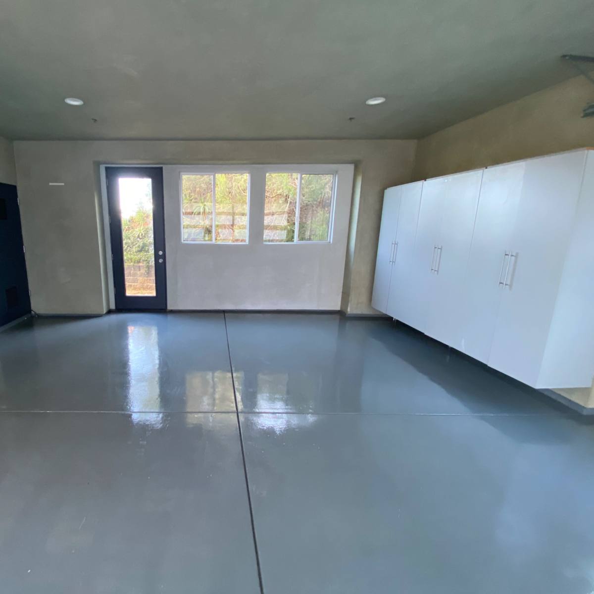 A vacant room with gray floors, a large window, a door, and white cabinets along one wall.