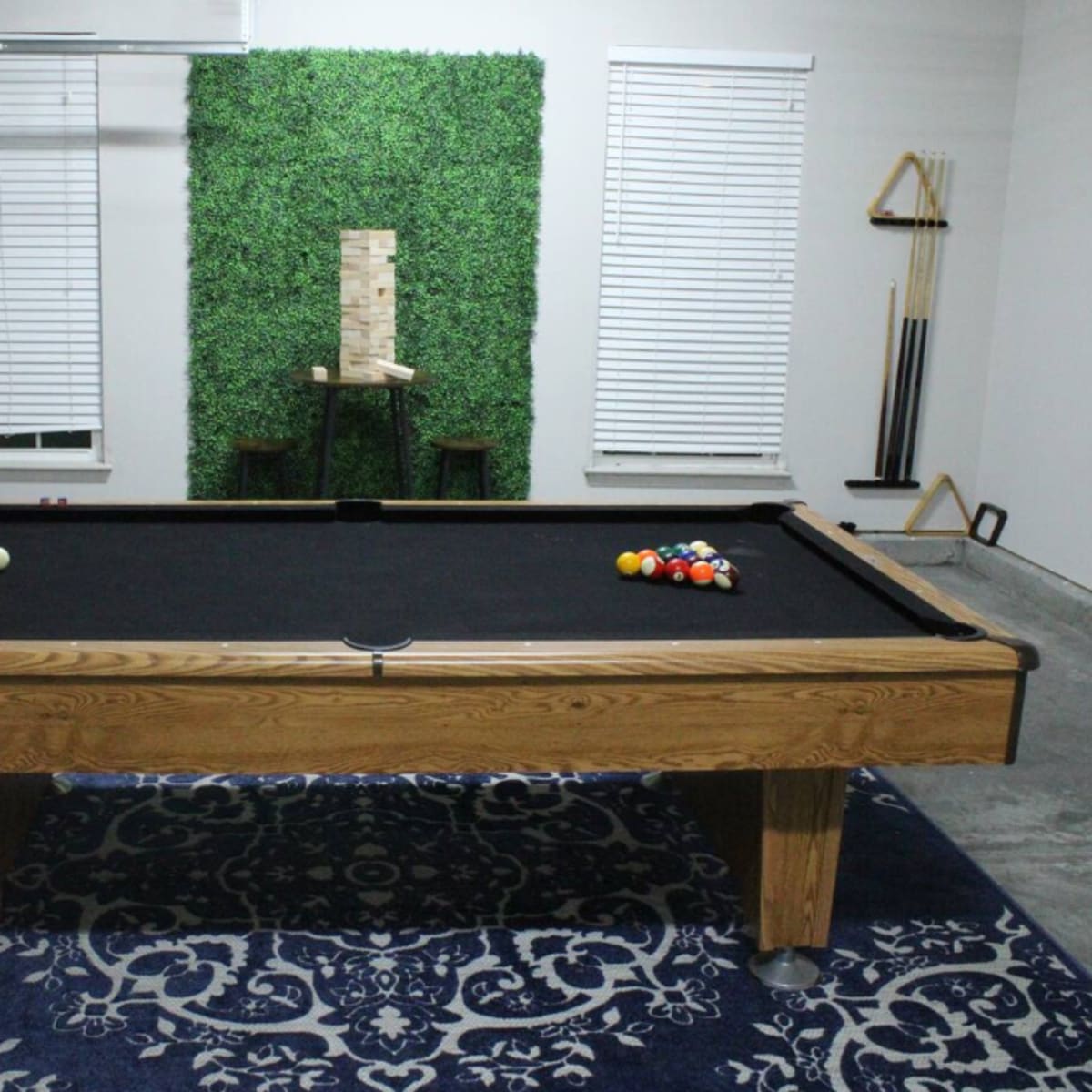 A billiards table with colorful balls set up on it is positioned in a garage-like space featuring a green wall, wooden decor, and window blinds.