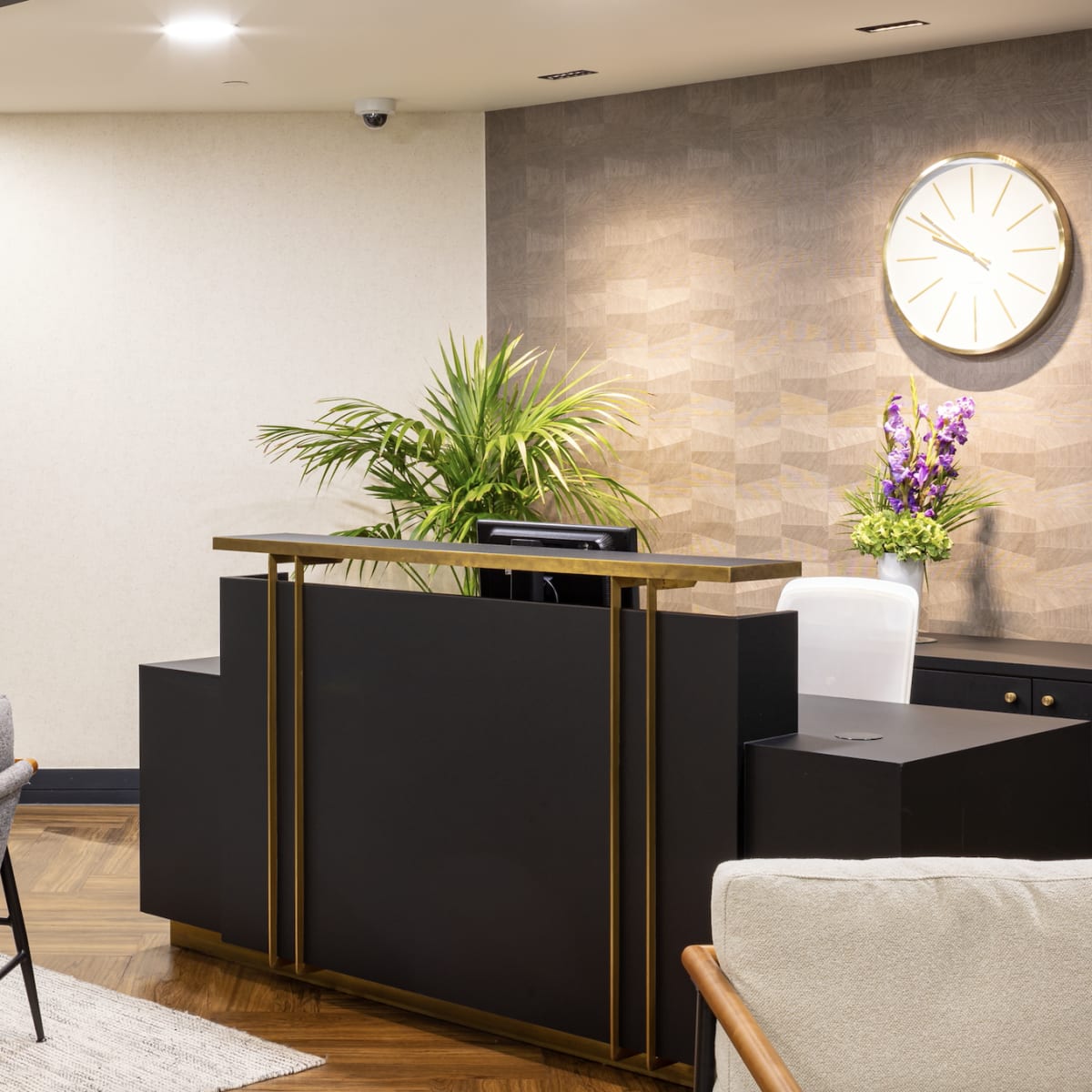 A modern reception area features a dark wooden front desk, a wall clock, and seating arrangements with armchairs and a rug.