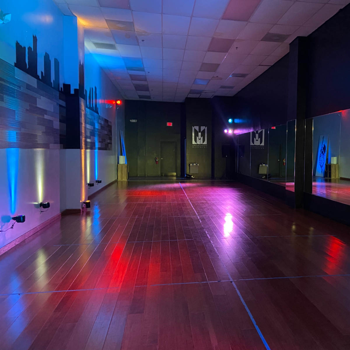 An empty dance studio with colorful lighting and mirrors reflecting a hardwood floor.