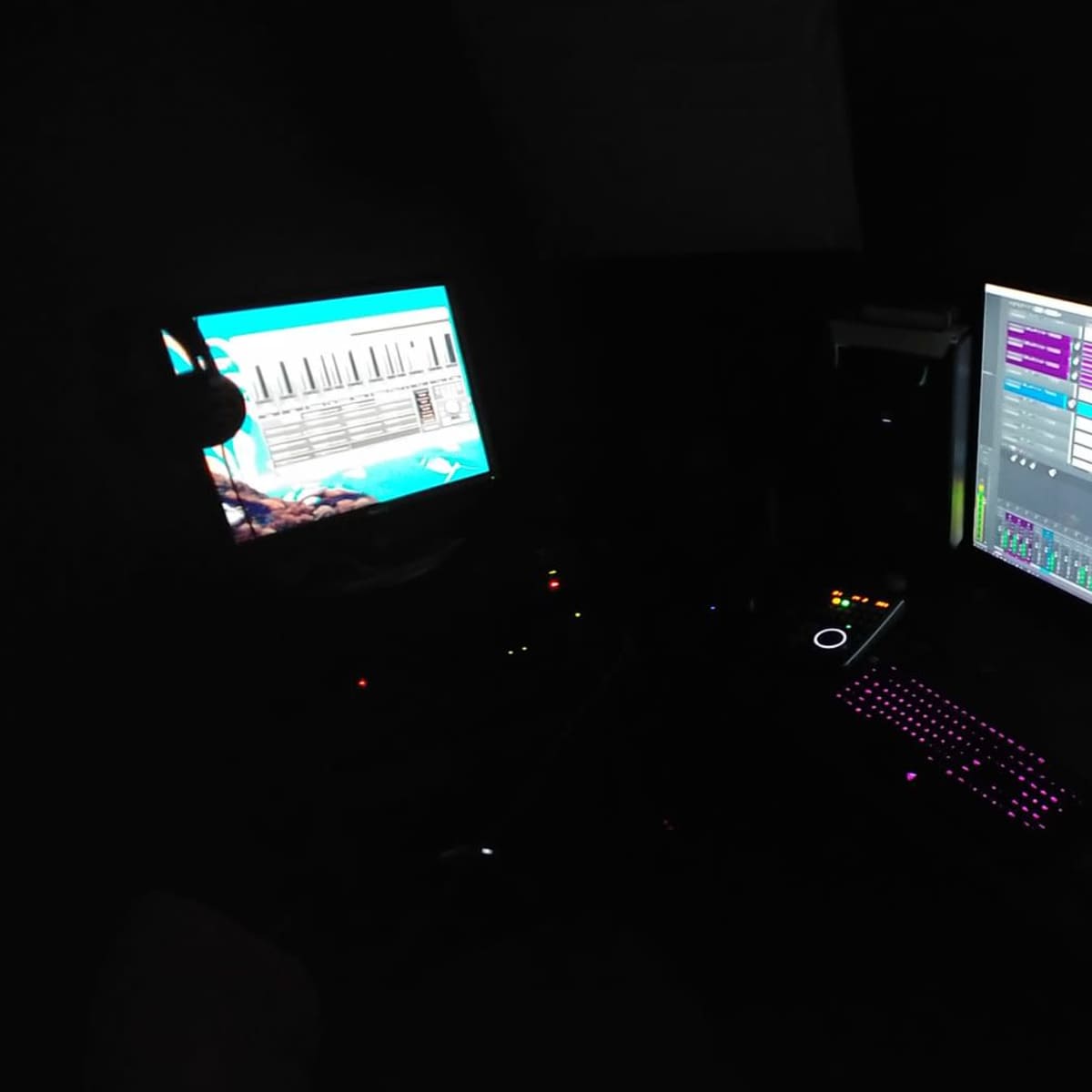 A dimly lit recording studio features two computer monitors displaying audio editing software and sound levels, surrounded by various equipment.