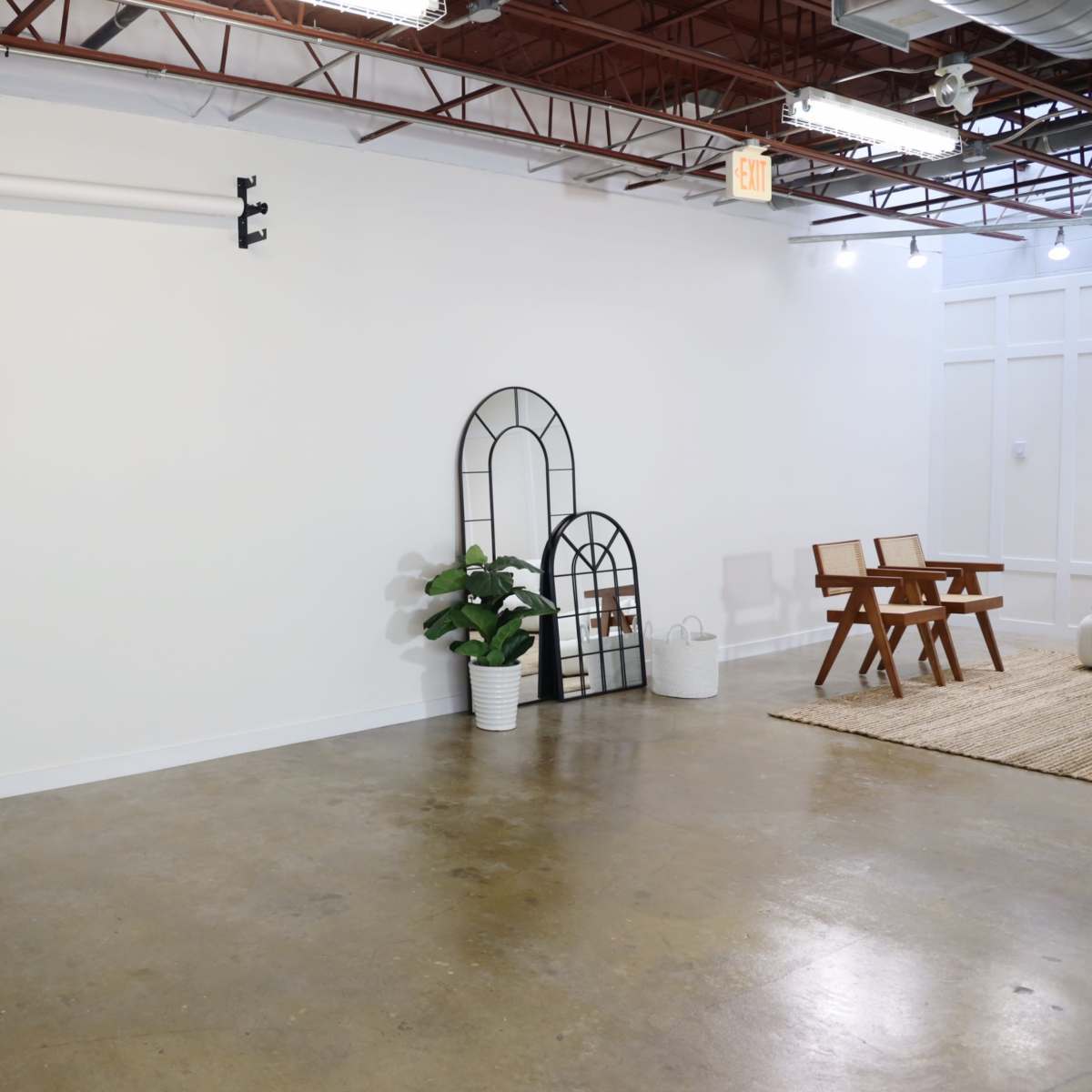 A spacious, empty room with a concrete floor, a few decorative elements including two arched mirrors and a potted plant, and a pair of wooden chairs on a woven rug.