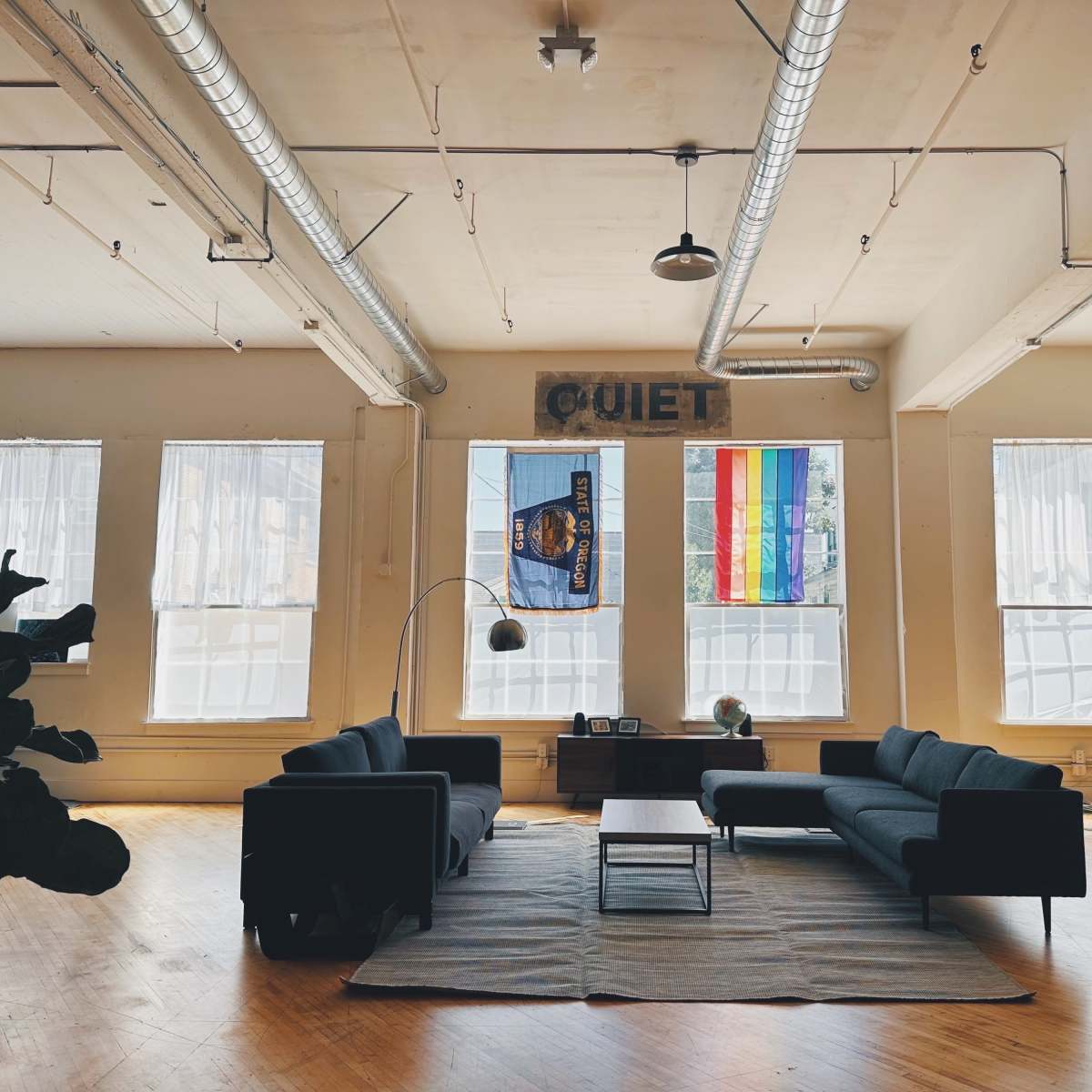 A spacious room with two large windows, featuring a minimalist seating area with a black sofa, a round lamp, and a rug on a wooden floor, along with colorful flags hanging in the windows.