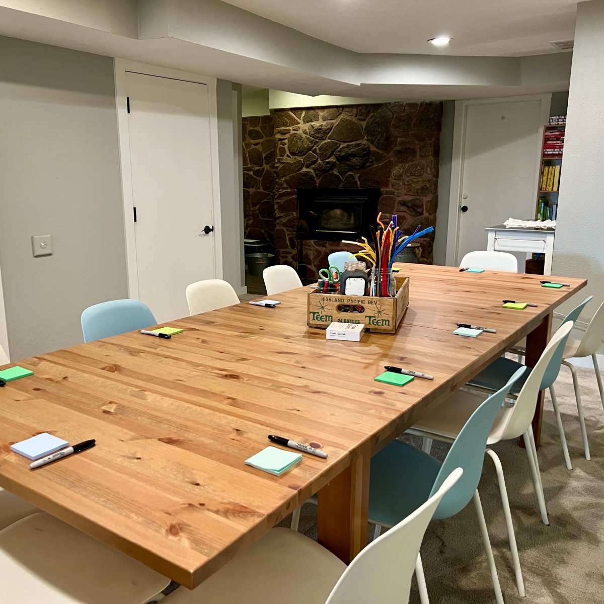 A conference room features a large wooden table surrounded by chairs, with a variety of stationery on the surface and a stone fireplace in the background.