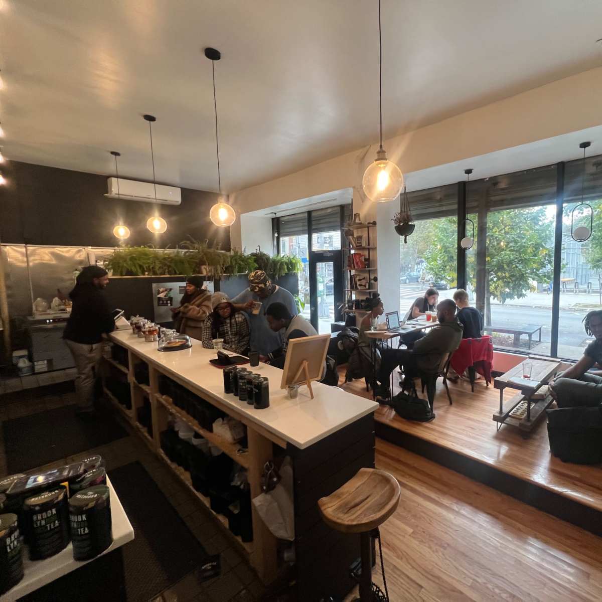 A cozy café features a wooden counter, several patrons seated at tables, and large windows showcasing an outdoor view.