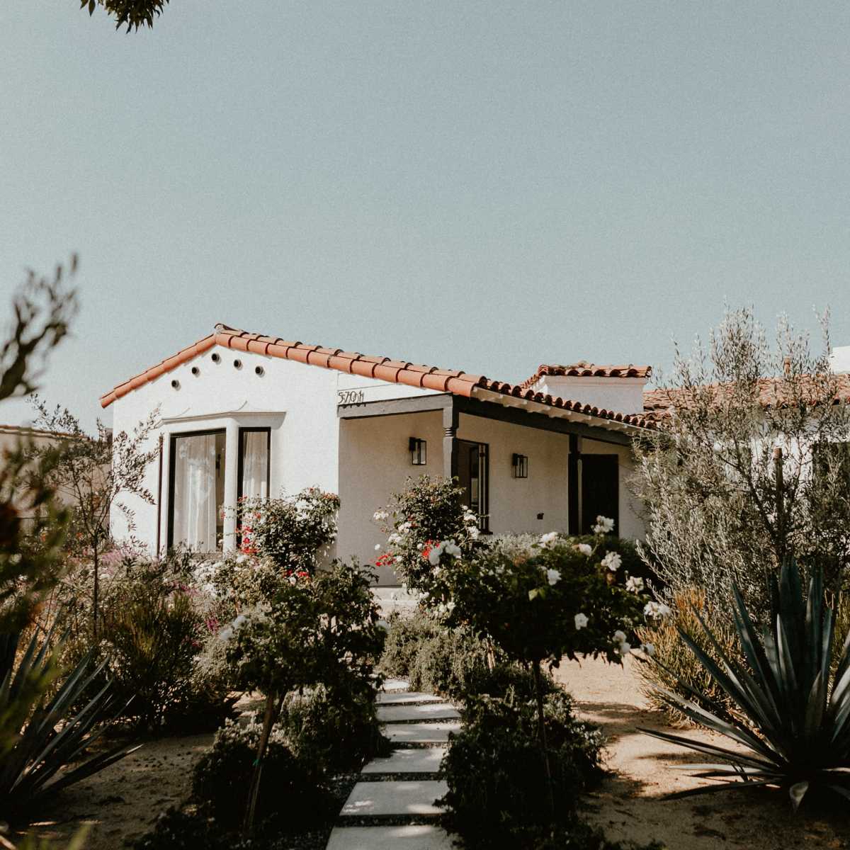 A single-story white house with a red-tiled roof is surrounded by desert landscaping and flowering shrubs.