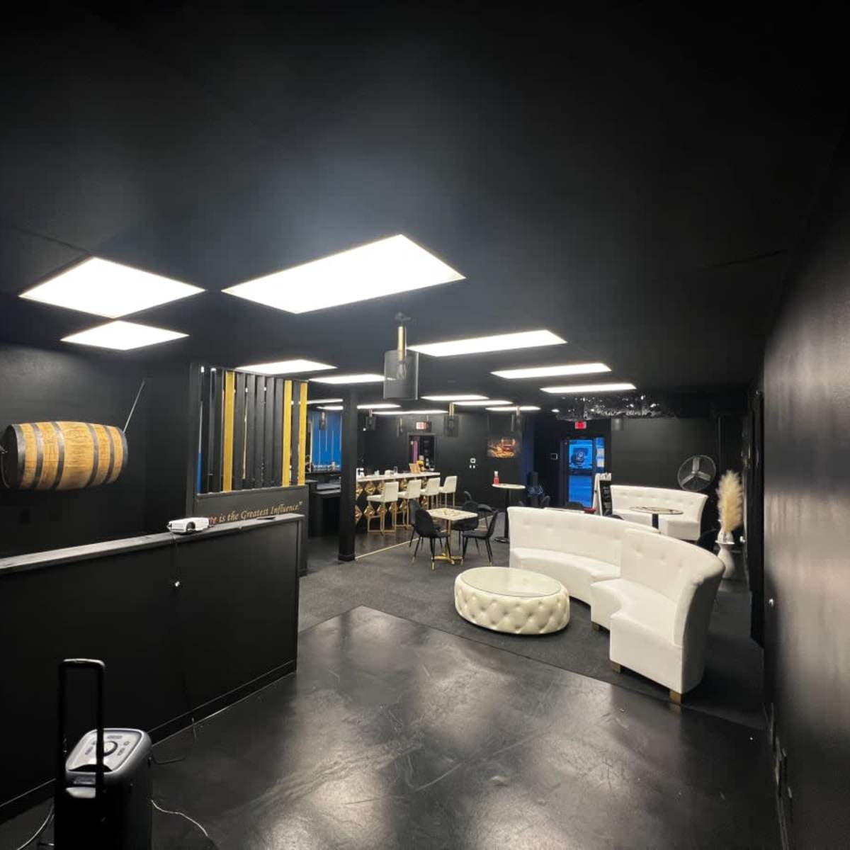 A modern black-walled room featuring a seating area with white couches, a dining area with a long table, and a wooden barrel hanging on the wall.
