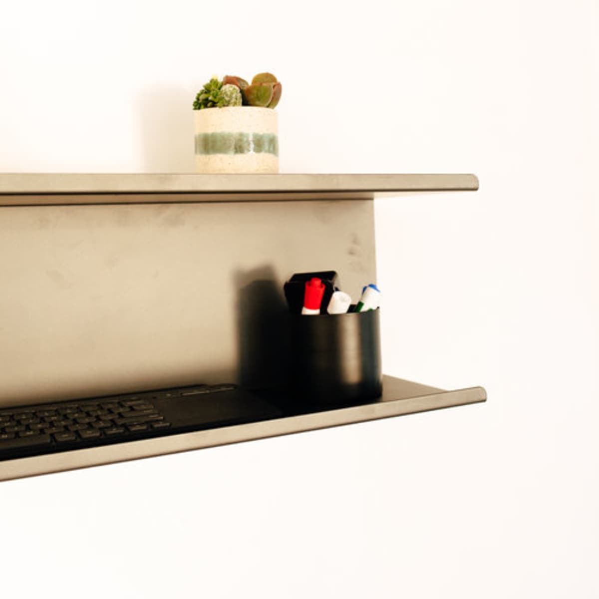 A black shelf holds a small potted plant and a container with various writing instruments, positioned against a white wall.