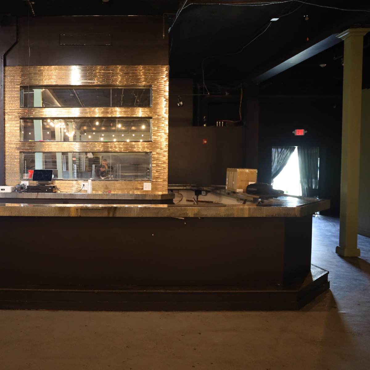 An empty bar with a polished countertop and a large mirror backdrop is shown in a dimly lit venue.