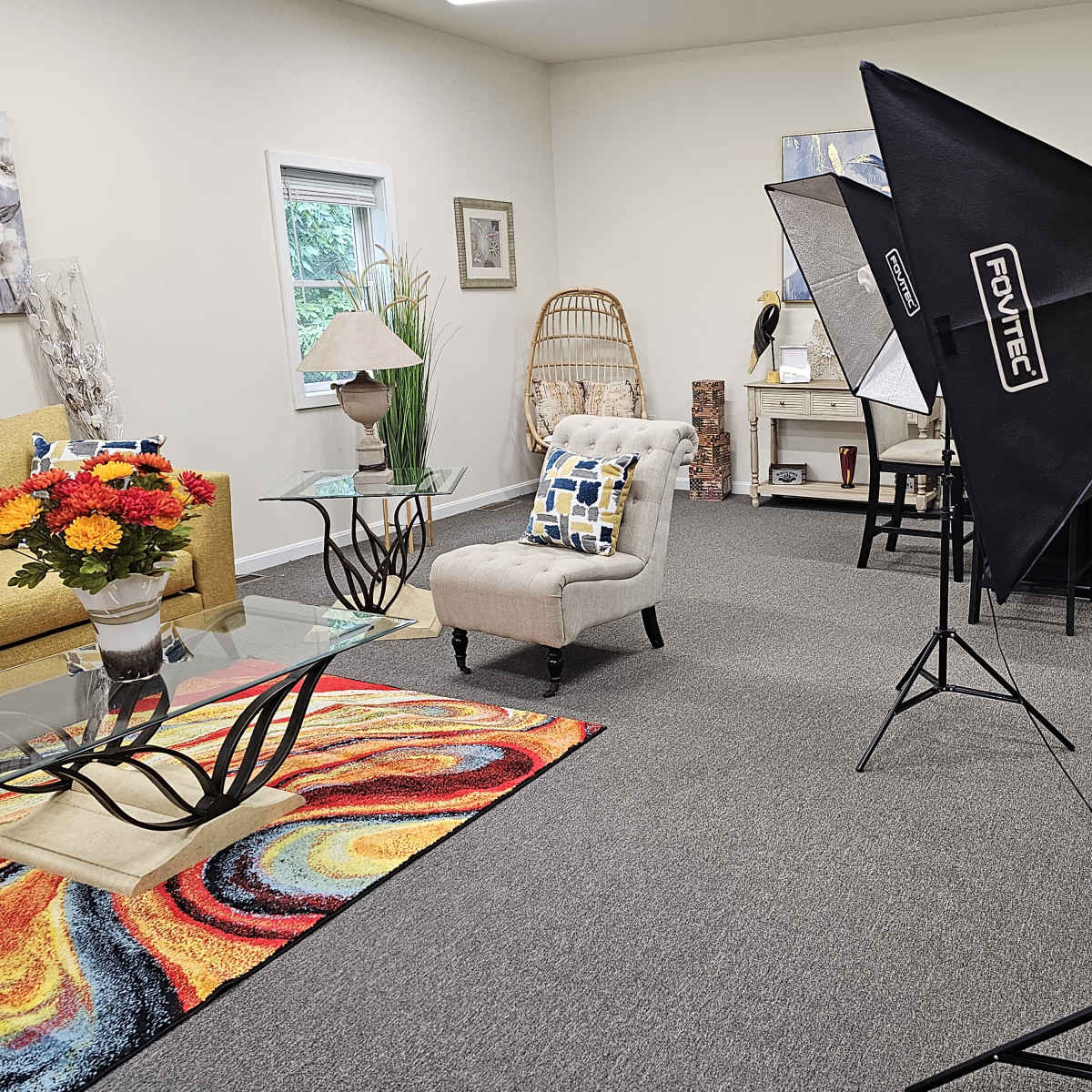A brightly lit room features a seating area with a yellow sofa, a glass coffee table on a colorful rug, and a corner with a lamp and artwork.
