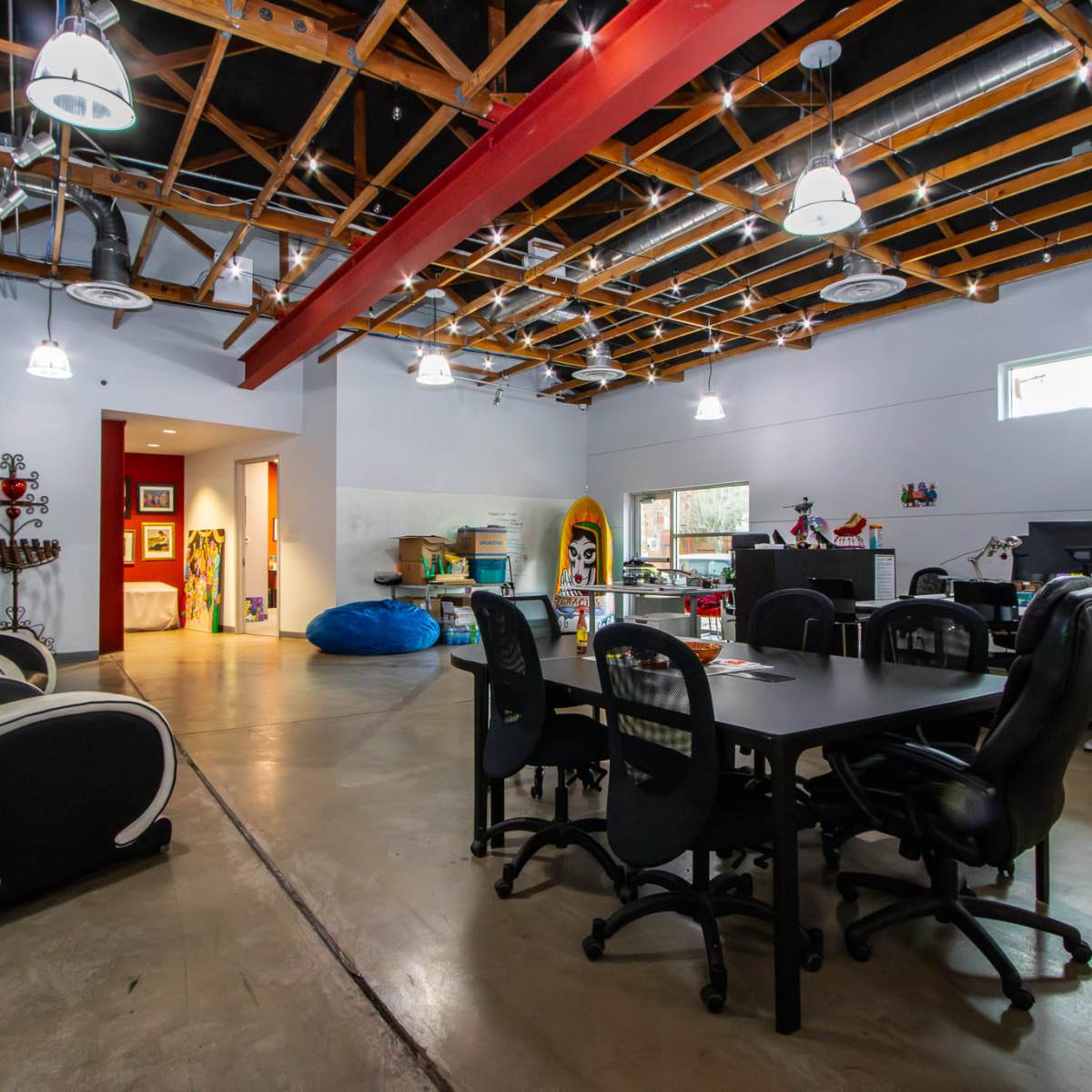 A spacious office area with a concrete floor, large windows, and a wood-beamed ceiling, featuring several desks, chairs, and a blue bean bag.