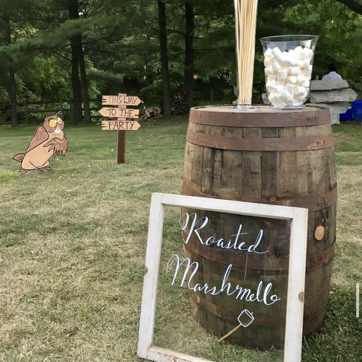 A wooden barrel displays a sign reading "Roasted Marshmelloe" alongside a glass container filled with marshmallows, while a whimsical sign points guests towards the party.