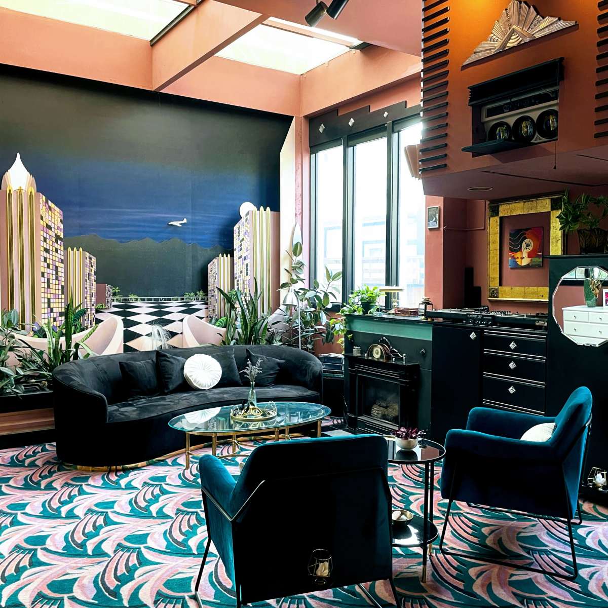 The scene features a stylish lounge area with a dark-colored couch and armchairs, surrounded by large windows and decorated with potted plants and a vibrant patterned carpet.