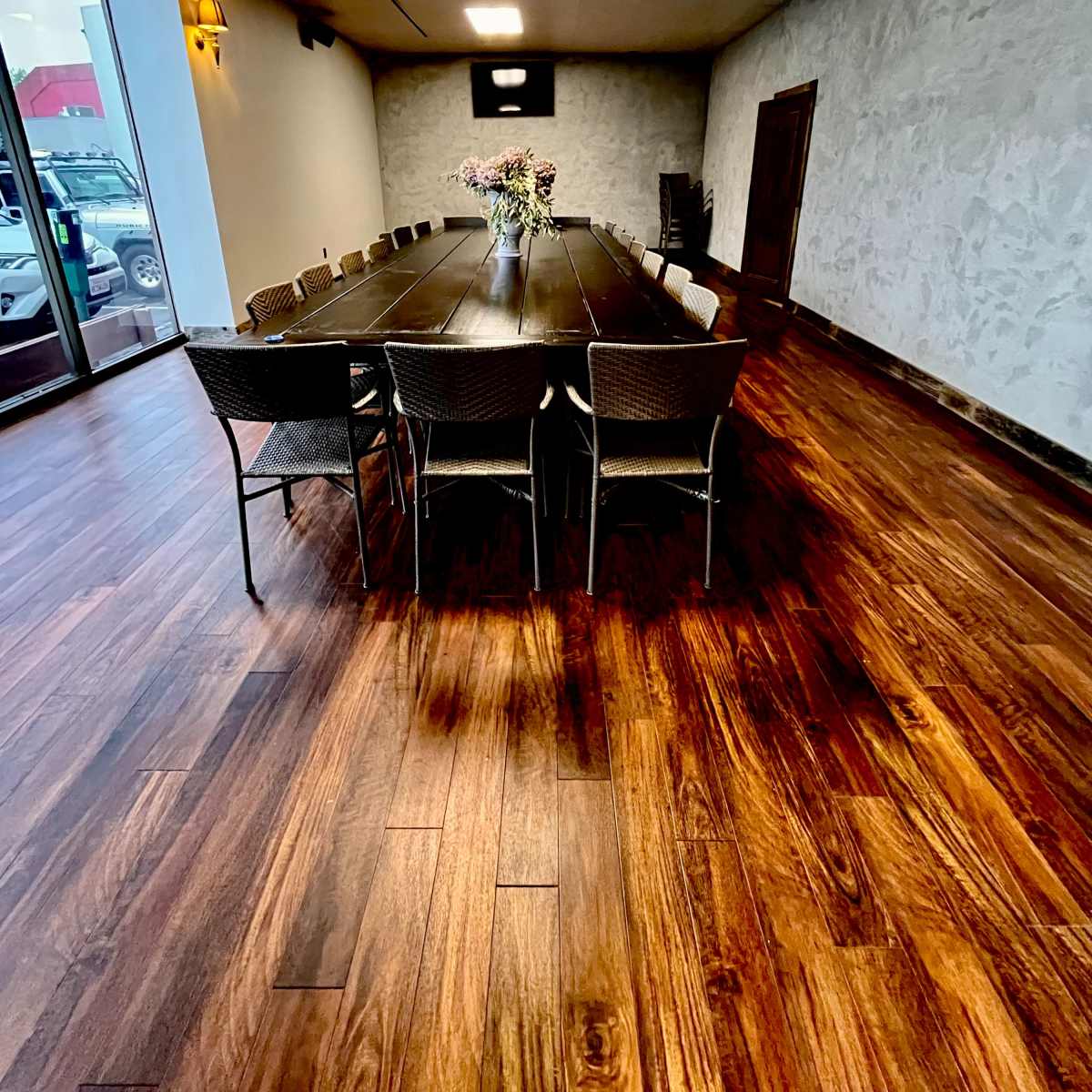 A long wooden table surrounded by chairs is positioned in a spacious room with large windows and wooden flooring.