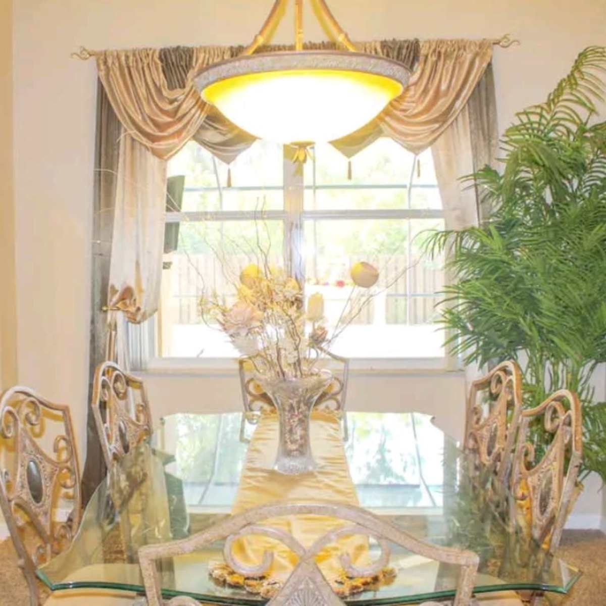 A dining room features a glass table surrounded by six ornate chairs, with a large light fixture above and a potted plant in the corner.