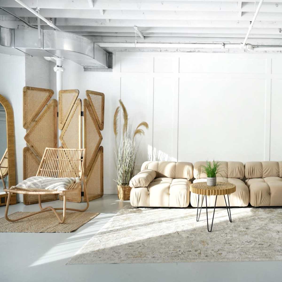 A spacious, light-filled room featuring a beige sectional sofa, a round wooden coffee table, a soft rug, and a rattan chair next to decorative plants.