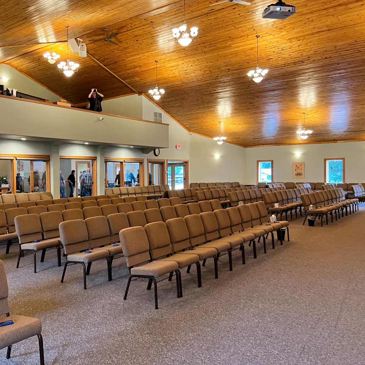 A spacious indoor venue with rows of brown chairs facing a stage area, illuminated by overhead lights and a wooden ceiling.