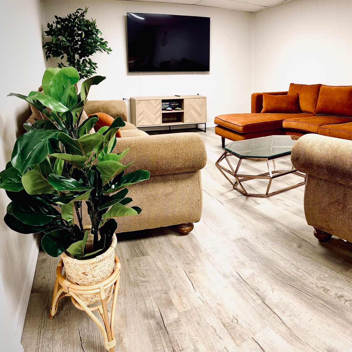 A modern, low-lit living room features a large TV mounted on the wall, a glass coffee table, and an arrangement of orange and brown sofas with a decorative plant in a wicker pot.