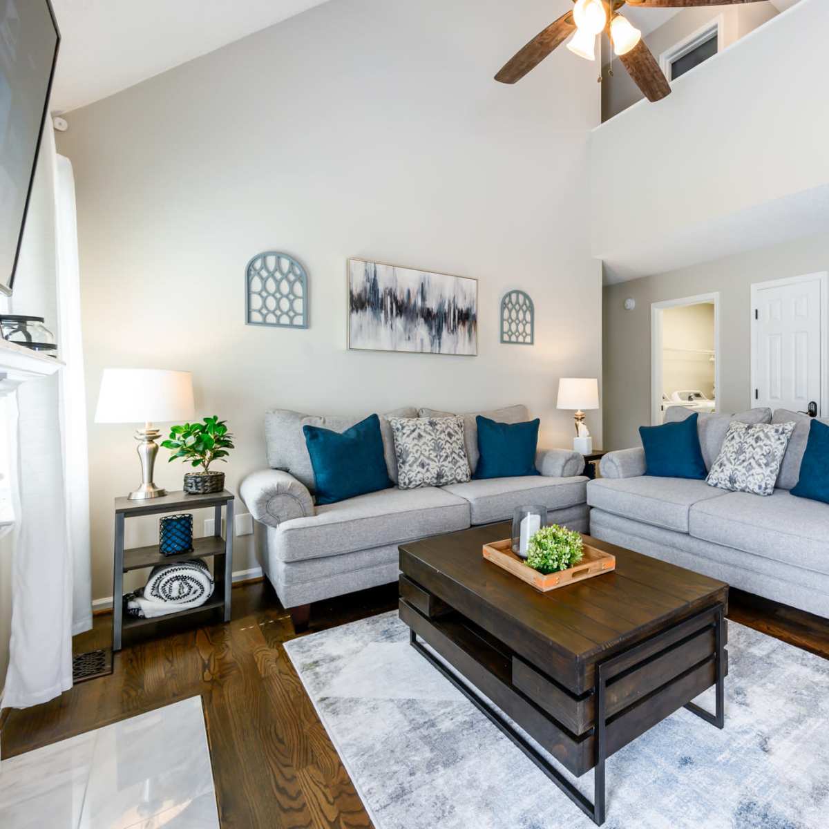 A living room features two gray sofas with blue accent pillows facing a dark wood coffee table on a light area rug, with a staircase and entrance visible in the background.