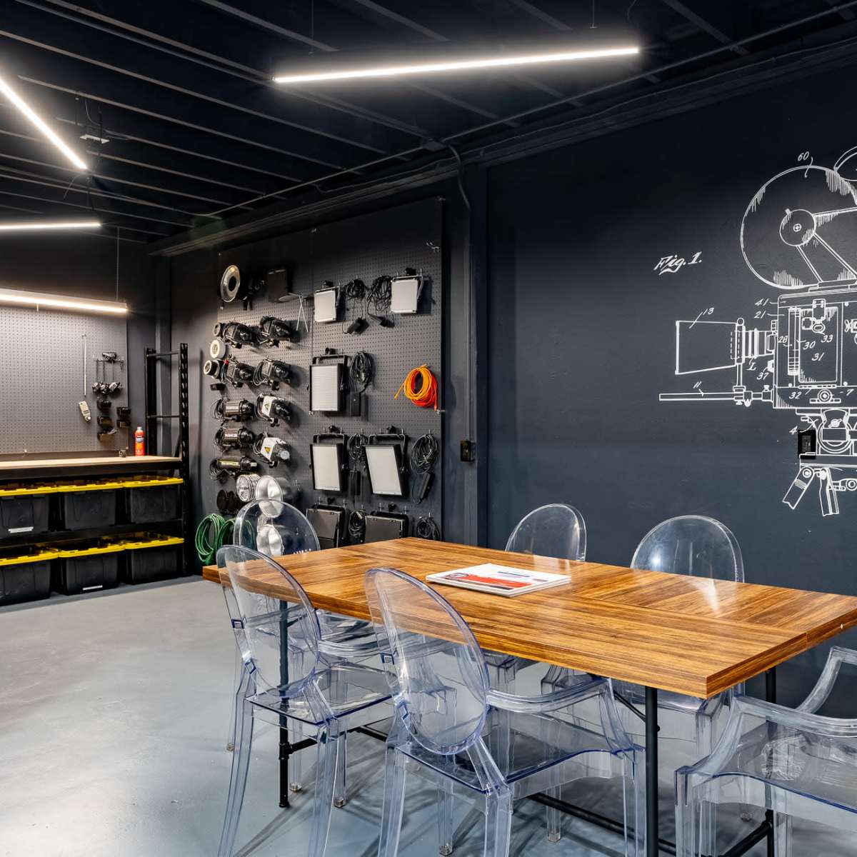 A modern workshop with a long wooden table surrounded by transparent chairs, and walls adorned with tools and a large illustration of a film camera.