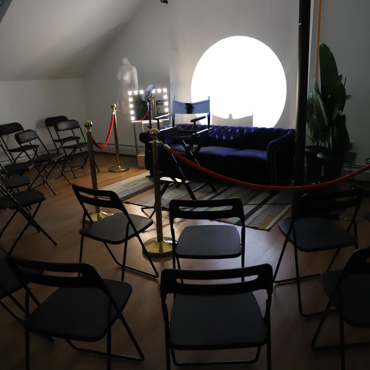 A room set up with black folding chairs arranged around a blue couch, accompanied by a spotlight on a white wall and red ropes creating a barrier.