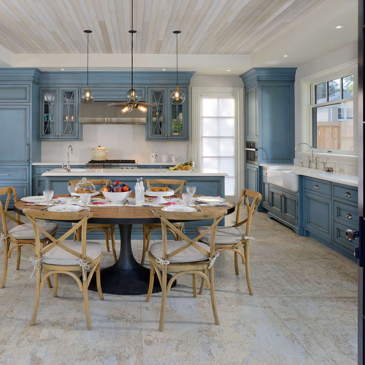 A modern kitchen features blue cabinetry, a large wooden dining table set with plates and food, and large windows providing natural light.