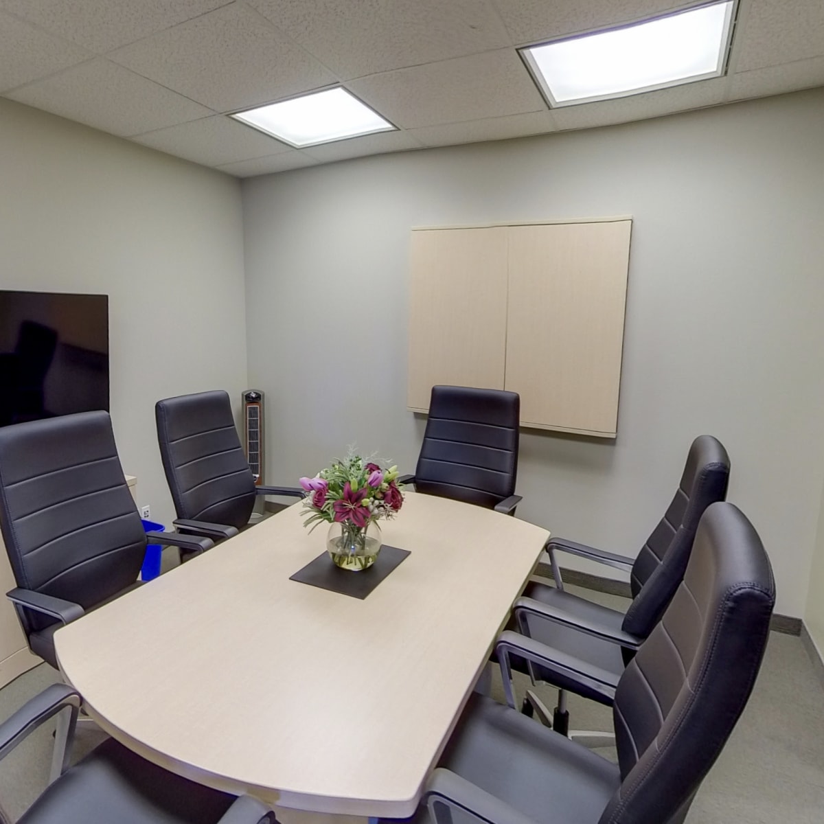 A conference room is set up with a large table surrounded by six black chairs, a wall-mounted screen, and a flower arrangement in the center.