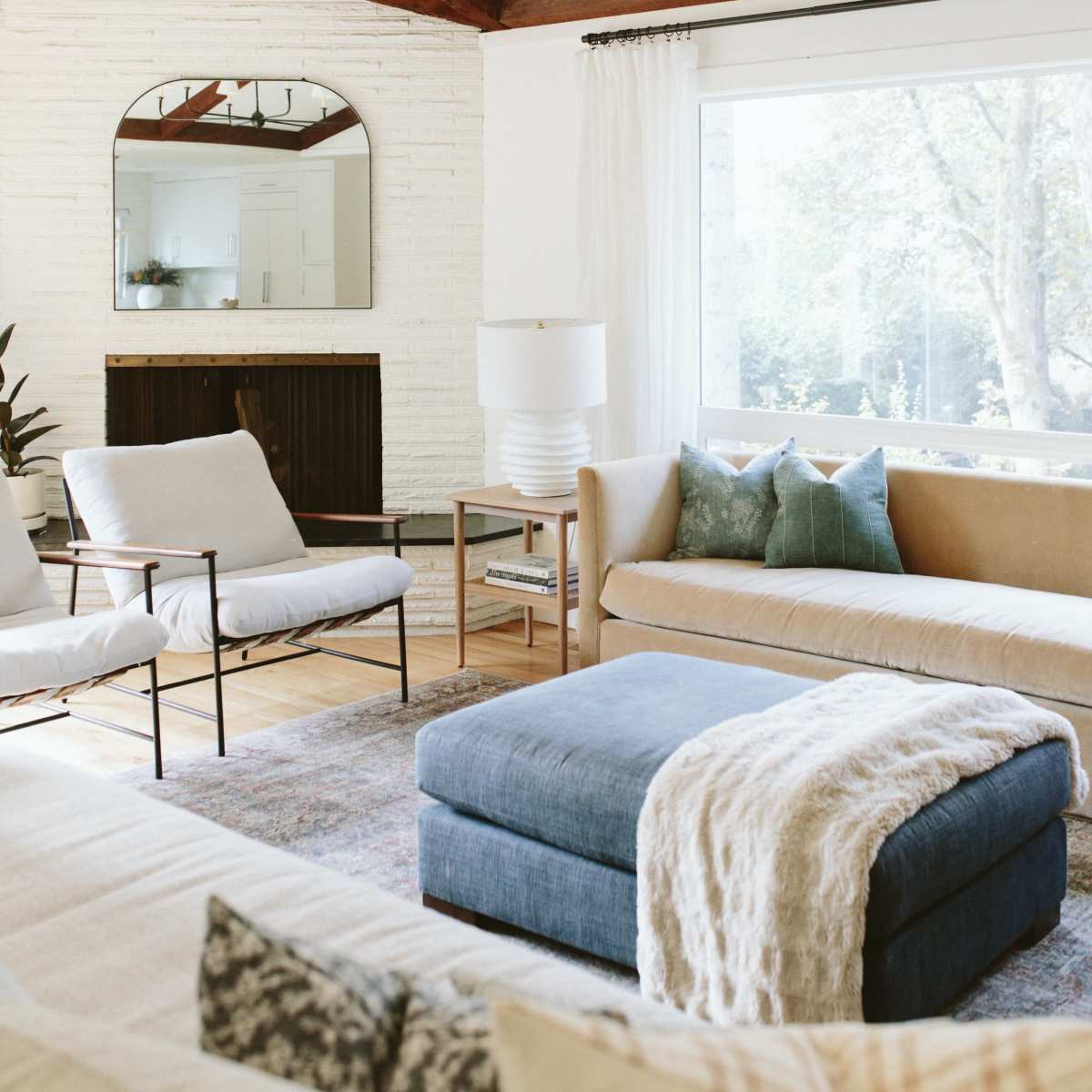 The living room features a light-colored sofa, a blue ottoman, and two armchairs, with large windows allowing natural light to fill the space, complemented by wooden beams overhead.