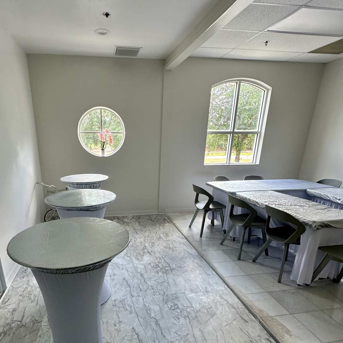A room with two round tables and a rectangular table, featuring large windows that allow natural light to enter.