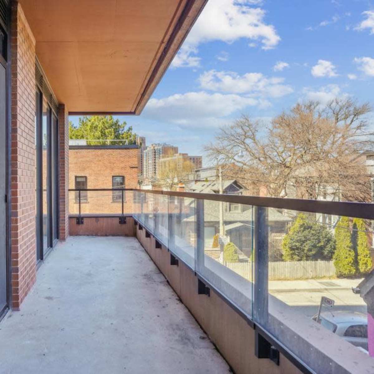 A wide balcony with glass railings overlooks a neighborhood with trees and buildings under a partly cloudy sky.